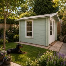 Exterior view of the Shire Barnsdale log cabin painted in a light sage green finish, set in a garden with a flagstone patio and surrounding trees.